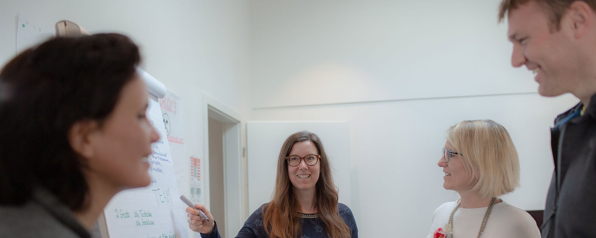 gute Stimmung: Drei Frauen und ein Mann stehen um ein Flipchart. Eine Frau mit langen Haaren und Brille zeigt mit dem Marker auf eine Stelle mit dem Wort "TEAM"