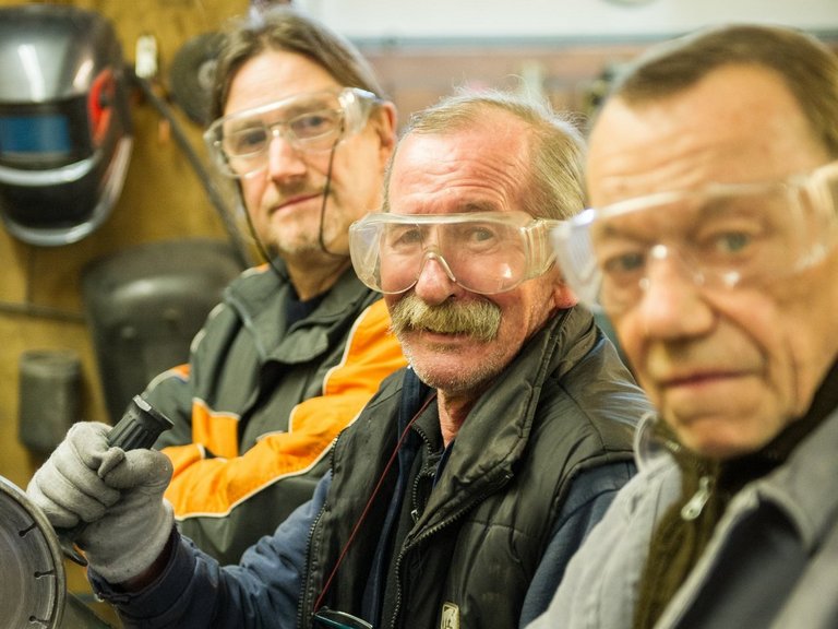 drei Männer mit Schutzbrille blicken in die Kamera - einer hält eine Flex in den Händen