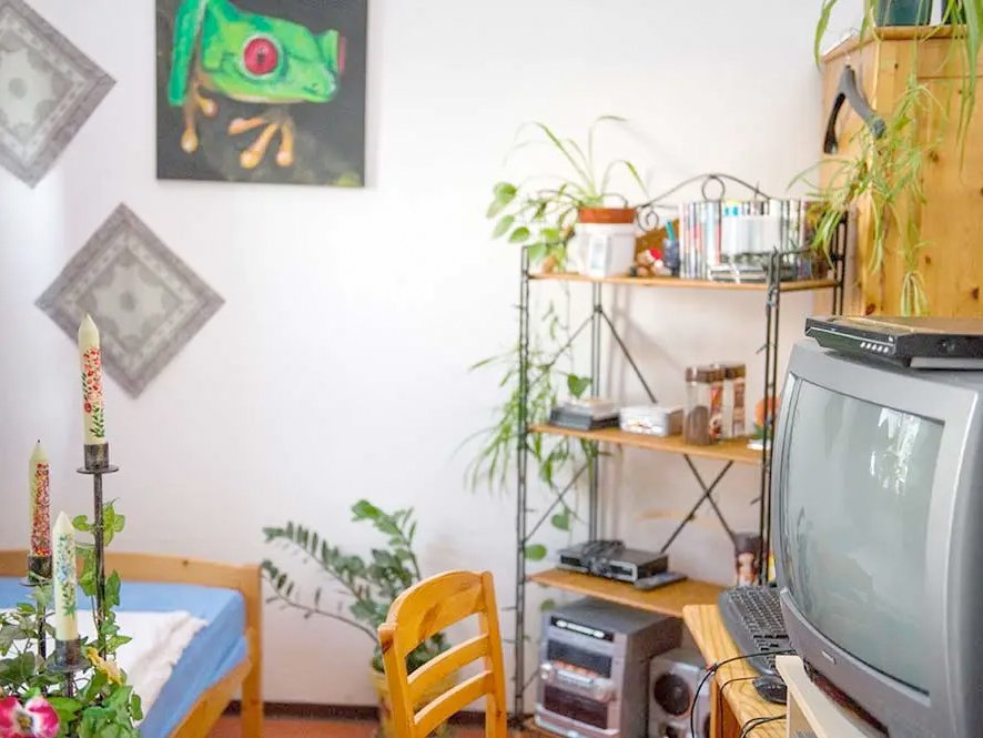 Gemütliches Zimmer mit Bett, Schrank und Regal. Im Vordergrund ein Fernseher und Kerzen. Dahinter Gemälde (ein grüner Frosch,...) an der Wand und die Stereoanlage im Regal.