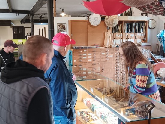 Besucher stehen vor einer Glasvitrine in einem Museum. Eine Frau mit buntem Pullover hinter der Vitrine zeigt ihnen Exponate. Im Hintergrund hängen bunte Regenschirme von der Decke und es stehen Holzschubladenschränke.