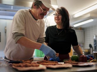 Eine Frau und ein lächelnder Mann mit Mütze, Brille und blauen Handschuhen bereiten eine Wurstplatte auf einem Edelstahltablett in einer Großküche zu. Im Hintergrund Küchengeräte unscharf zu erkennen.