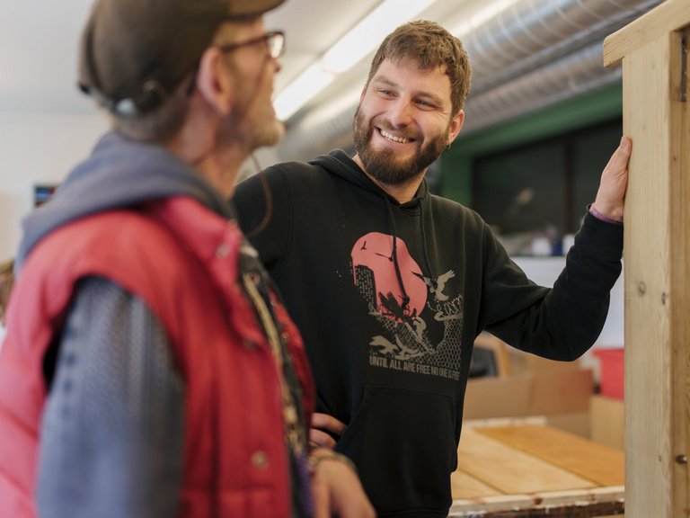 Zwei Männer unterhalten sich in einer Holzwerkstatt. Ein bärtiger Mann in einem schwarzen Hoodie lächelt und stützt sich an einem hölzernen Regal ab, während ihm ein Mann in roter Weste zuhört.