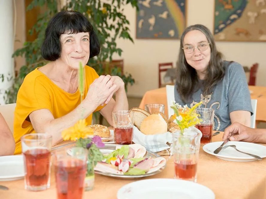 zwei glücklich wirkende Frauen sitzen am Frühstückstisch bei Brot, Brötchen und schön garniertem Wurstaufschnitt