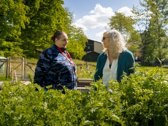 Zwei Frauen unterhalten sich in einem sonnigen Garten, umgeben von üppigem Grün. Die Frau links trägt eine blaue Jacke, die Frau rechts einen türkisfarbenen Blazer. Im Hintergrund sind Bäume und ein Haus zu erkennen.