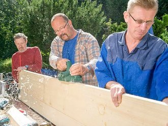 Zwei Männer halten eine Holzplatte. Ein weiterer hobelt mit der Maschine eine Seite ab.