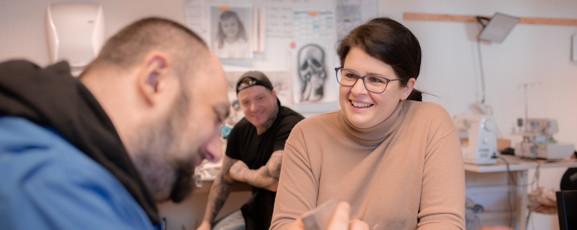 Eine freundliche Mitarbeiterin mit Brille und dunklen Haaren ist im Gespräch mit einem jungen Mann, der scheinbar gerade an einem Werkstück arbeitet