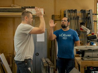 Zwei Männer in einer Holzwerkstatt geben sich ein High-Five. Der Mann links trägt ein weißes T-Shirt, der Mann rechts ein blaues T-Shirt. Im Hintergrund sind Werkzeuge und Holzplanken zu sehen.