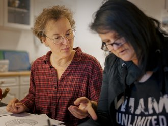 Zwei Frauen sitzen an einem Tisch und diskutieren über eine Zeichnung. Die Frau rechts im schwarzen Hoodie hört zu, während die Frau links im karierten Hemd mit den Händen etwas erklärt. Beide tragen Brillen.